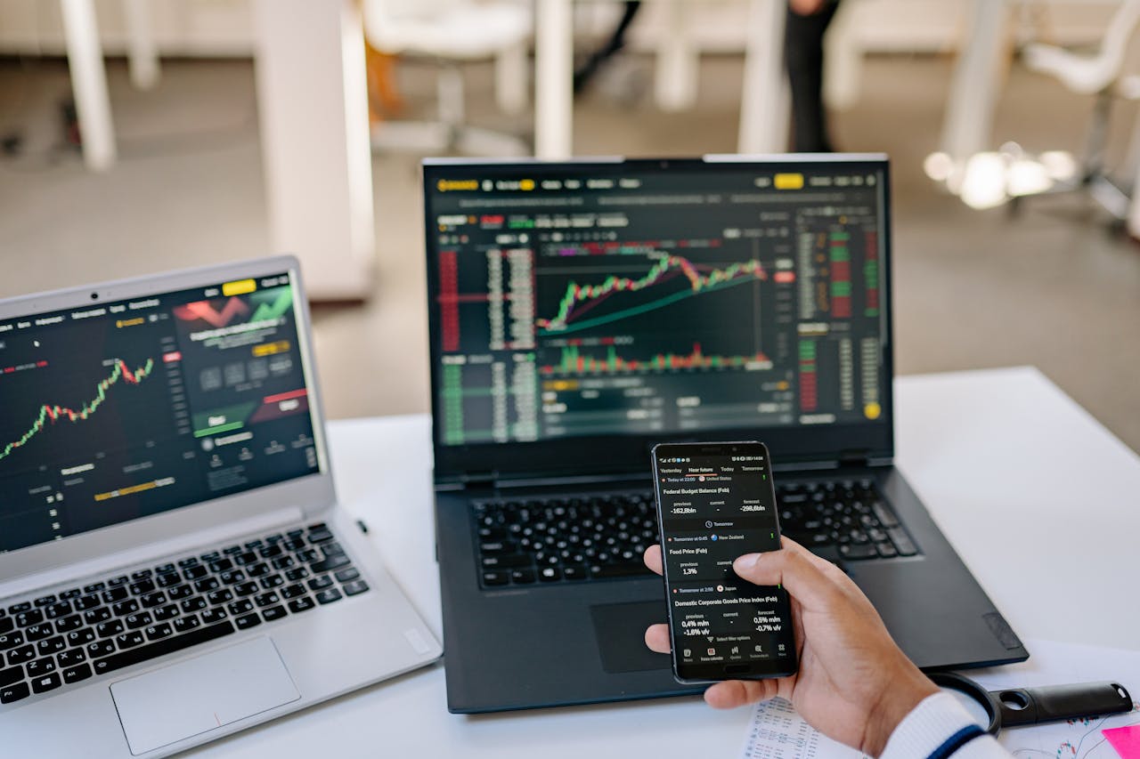 gallery-03 A professional tracks stock market trends using two laptops and a smartphone, showcasing digital finance tools.